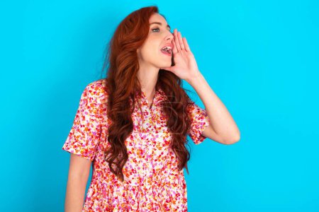 Photo for Redhead woman wearing floral dress over blue background profile view, looking happy and excited, shouting and calling to copy space. - Royalty Free Image