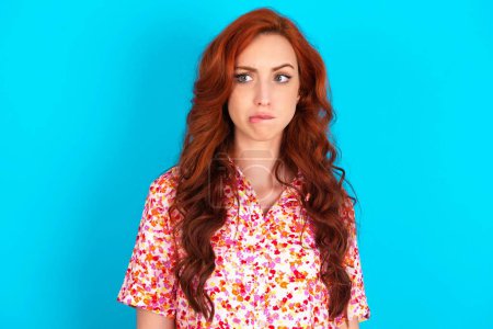 Photo for Redhead woman wearing floral dress over blue background being nervous and scared biting lips looking camera with impatient expression, pensive. - Royalty Free Image