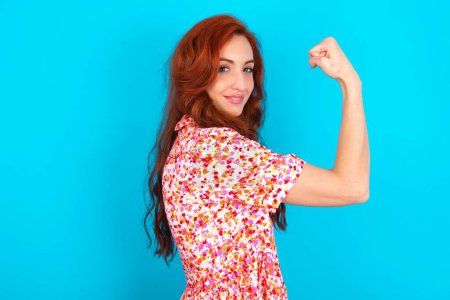 Photo for Redhead woman wearing floral dress over blue background showing muscles after workout. Health and strength concept. - Royalty Free Image