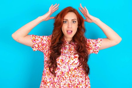 Photo for Redhead woman wearing floral dress over blue background looks with excitement at camera, keeps hands raised over head, notices something unexpected, reacts on sudden news. - Royalty Free Image