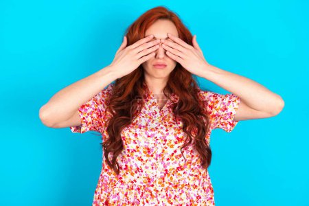 Photo for Redhead woman wearing floral dress over blue background covering eyes with both hands, doesn't want to see anything or feeling ashamed. Human feelings reactions. - Royalty Free Image