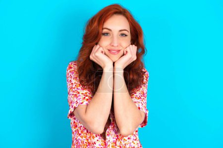 Photo for Dreamy redhead woman wearing floral dress over blue background keeps hands pressed together under chin, looks with happy expression, has toothy smile. - Royalty Free Image