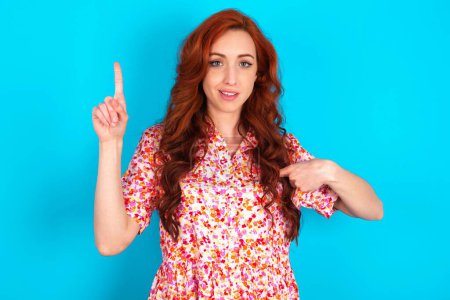 Photo for Redhead woman wearing floral dress over blue background  says: wow how exciting it is, has amazed expression, indicates something. One hand on her chest and pointing with other hand. - Royalty Free Image