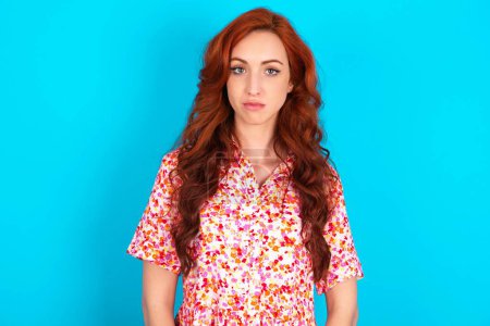 Photo for Joyful redhead woman wearing floral dress over blue background looking to the camera, thinking about something. Both arms down, neutral facial expression. - Royalty Free Image