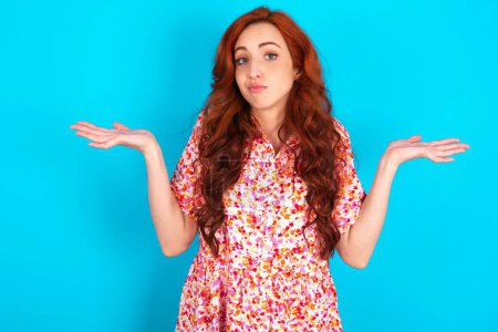 Photo for Puzzled and clueless redhead woman wearing floral dress over blue background with arms out, shrugging shoulders, saying: who cares, so what, I don't know. - Royalty Free Image