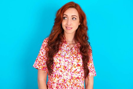 Photo for Oops! Portrait of redhead woman wearing floral dress over blue background clenches teeth and looks confusedly aside, realizes her bad mistake, - Royalty Free Image