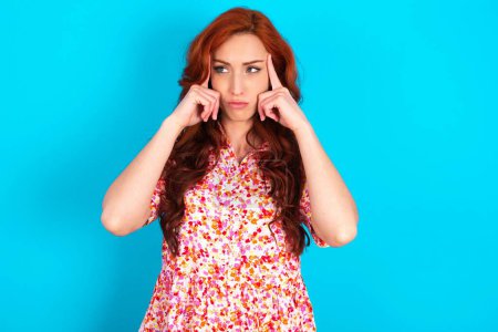 Photo for Redhead woman wearing floral dress over blue background with thoughtful expression, looks away, keeps hand near face, thinks about something pleasant. - Royalty Free Image