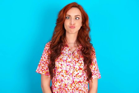 Photo for Redhead woman wearing floral dress over blue background crosses eyes, puts lips, makes grimace with awkward expression has fun alone, plays fool. - Royalty Free Image