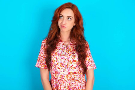 Photo for Redhead woman wearing floral dress over blue background has worried face looking up lips together, being upset thinking about something important, keeps hands down. - Royalty Free Image