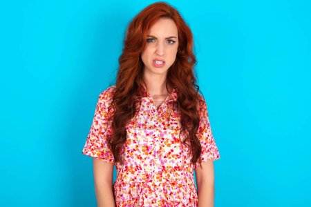 Photo for Mad crazy redhead woman wearing floral dress over blue background clenches teeth angrily, being annoyed with coming noise. Negative feeling concept. - Royalty Free Image