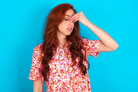 Photo for Very upset, redhead woman wearing floral dress over blue background touching nose between closed eyes, wants to cry, having stressful relationship or having troubles with work - Royalty Free Image