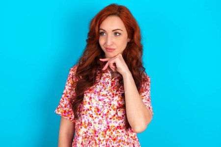 Photo for Dreamy redhead woman wearing floral dress over blue background with pleasant expression, looks sideways, keeps hand under chin, thinks about something pleasant. - Royalty Free Image