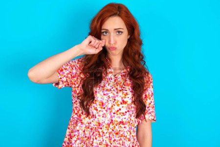 Photo for Unhappy redhead woman wearing floral dress over blue background crying while posing at camera whipping tears with hand. - Royalty Free Image