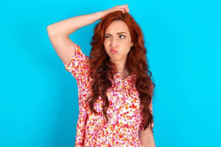 Photo for Redhead woman wearing floral dress over blue background saying: Oops, what did I do? Holding hand on head with frightened and regret expression. - Royalty Free Image
