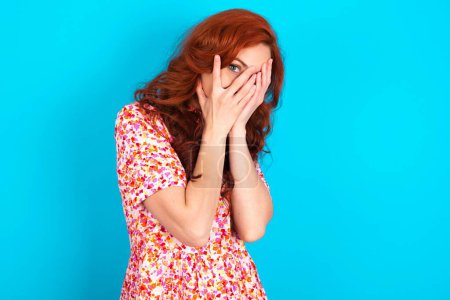 Photo for Redhead woman wearing floral dress over blue background covering face with hands and peering out with one eye between fingers. Scared from something or someone. - Royalty Free Image