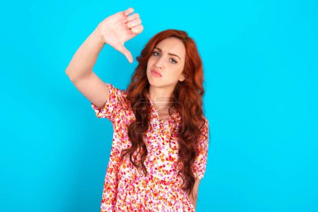 Photo for Discontent redhead woman wearing floral dress over blue background shows disapproval sign, keeps thumb down, expresses dislike, frowns face in discontent. Negative feelings. - Royalty Free Image
