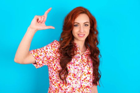Photo for Redhead woman wearing floral dress over blue background smiling and gesturing with hand small size, measure symbol. - Royalty Free Image