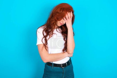 Photo for Redhead woman wearing white T-shirt over blue background making facepalm gesture while smiling amazed with stupid situation. - Royalty Free Image