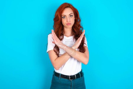 Photo for Redhead woman wearing white T-shirt over blue background Has rejection angry expression on face and crossing hands doing refusal negative sign. - Royalty Free Image