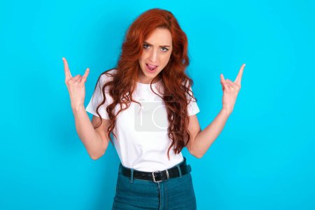 Photo for Born to rock this world. Joyful redhead woman wearing white T-shirt over blue background screaming out loud and showing with raised arms horns or rock gesture. - Royalty Free Image