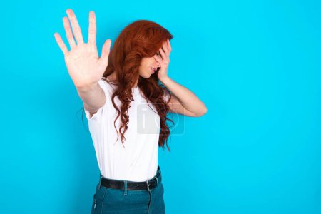 Photo for Redhead woman wearing white T-shirt over blue background covers eyes with palm and doing stop gesture, tries to hide. Don't look at me, I don't want to see, feels ashamed or scared. - Royalty Free Image