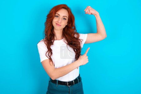 Téléchargez les photos : Femme rousse souriante portant un t-shirt blanc sur fond bleu lève la main pour montrer les muscles, se sent confiante dans la victoire, forte et indépendante. - en image libre de droit