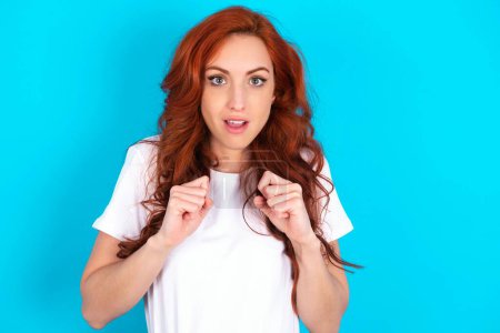 Photo for Portrait of desperate and shocked redhead woman wearing white T-shirt over blue background looking panic, holding hands near face, with mouth wide open. - Royalty Free Image