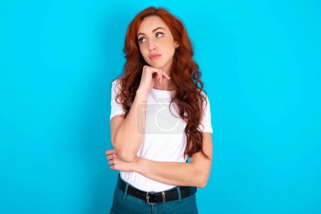 Face expressions and emotions. Thoughtful redhead woman wearing white T-shirt over blue background holding hand under his head, having doubtful look.
