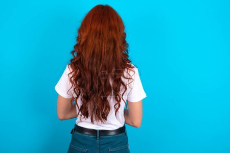Photo for Redhead woman wearing white T-shirt over blue background standing backwards looking away with arms on body. - Royalty Free Image