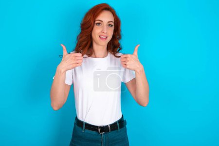 Foto de Mujer pelirroja optimista con camiseta blanca sobre fondo azul mostrando los pulgares hacia arriba con emociones positivas. Concepto de calidad y recomendación. - Imagen libre de derechos