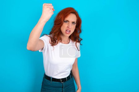 Foto de Mujer pelirroja feroz con camiseta blanca sobre fondo azul sosteniendo el puño delante como si estuviera lista para pelear o desafiar, gritando y teniendo expresión agresiva en la cara. - Imagen libre de derechos