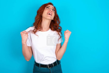 Téléchargez les photos : Rousse femme portant un t-shirt blanc sur fond bleu regarde avec excitation, garde les mains levées, remarque quelque chose d'inattendu. - en image libre de droit
