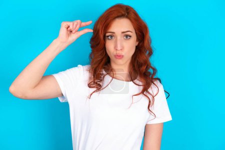Photo for Shocked redhead woman wearing white T-shirt over blue background shows something little with hands, demonstrates size, opens mouth from surprise. Measurement concept. - Royalty Free Image