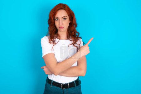 Photo for Redhead woman wearing white T-shirt over blue background smiling broadly at camera, pointing fingers away, showing something interesting and exciting. - Royalty Free Image