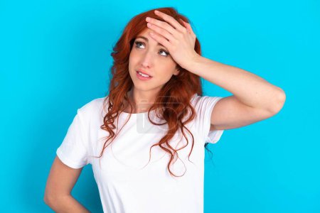 Photo for Redhead woman wearing white T-shirt over blue background touching forehead, hears something surprising, glad receive good news, feels relieved. Almost got in trouble. - Royalty Free Image
