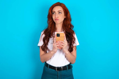 Photo for Portrait of redhead woman wearing white T-shirt over blue background with dreamy look, thinking while holding smartphone. Tries to write up a message. - Royalty Free Image