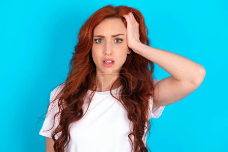Portrait of confused redhead woman wearing white T-shirt over blue background holding hand on hair and frowning, panicking, losing memory. Worried and anxious can not remember anything.