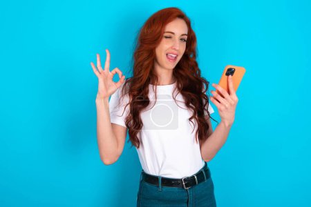 Téléchargez les photos : Joyeux rousse femme portant un t-shirt blanc envoyer un message sur son smartphone ou prendre un selfie et faire signe ok avec sa main. - en image libre de droit