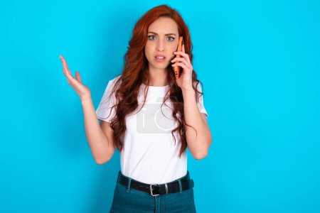 Photo for Redhead woman wearing white T-shirt talking on the phone stressed with hand on face, shocked with shame and surprise face, angry and frustrated. Fear and upset for mistake. - Royalty Free Image