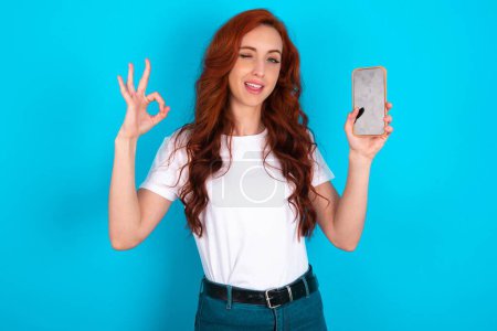 Foto de Mujer pelirroja emocionada vistiendo una camiseta blanca que muestra la pantalla en blanco del teléfono inteligente, parpadeando y haciendo señal de OK con la mano. Concepto de publicidad. - Imagen libre de derechos