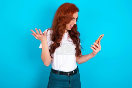 Photo for Photo of outraged annoyed redhead woman wearing white T-shirt holds cell phone, makes call, argues with colleague,  expresses negative emotions. People and anger. - Royalty Free Image