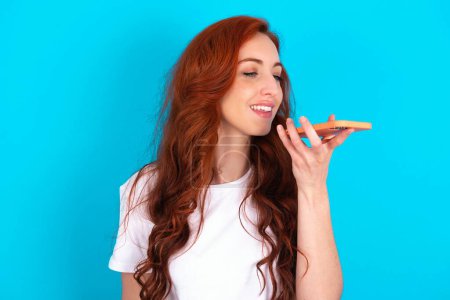 Photo for Smiling redhead woman wearing white T-shirt sending voice message on her smart phone. Communication and new technologies concept. - Royalty Free Image