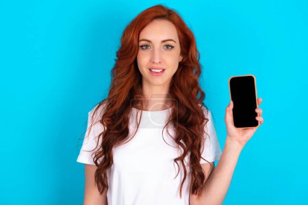 Téléchargez les photos : Femme rousse souriante portant un T-shirt blanc montrant un écran de téléphone vide. Concept de publicité et de communication. - en image libre de droit