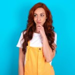 Thoughtful redhead woman wearing orange overall over blue background holds chin and looks away pensively makes up great plan