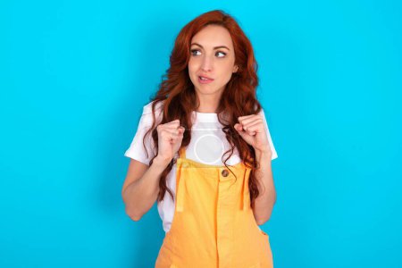 Photo for Redhead woman wearing orange overall over blue background clenches fists and awaits for something nice happened looks away bites lips and waits announcement of results - Royalty Free Image