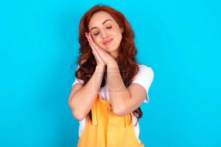 Photo for Redhead woman wearing orange overall over blue background leans on pressed palms closes eyes and has pleasant smile dreams about something - Royalty Free Image