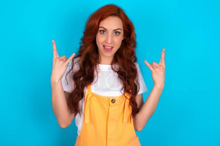 Photo for Redhead woman wearing orange overall over blue background makes rock n roll sign looks self confident and cheerful enjoys cool music at party. Body language concept. - Royalty Free Image