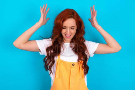 Photo for Redhead woman wearing orange overall over blue background goes crazy as head goes around feels stressed because of horrible - Royalty Free Image