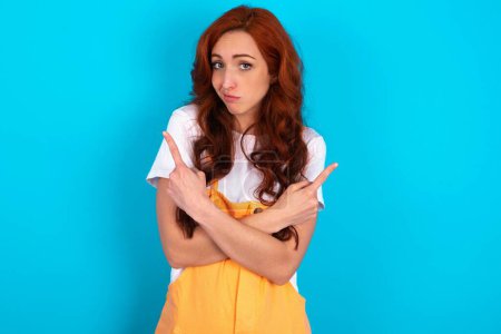 Photo for Redhead woman wearing orange overall over blue background crosses arms and points at different sides hesitates between two items or variants. Needs help with decision - Royalty Free Image
