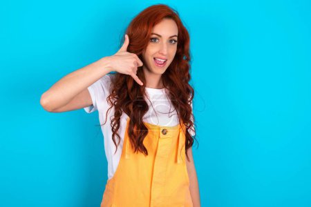 Photo for Redhead woman wearing orange overall over blue background makes phone gesture, says call me back again, has glad expression. - Royalty Free Image